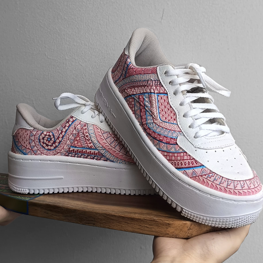 Hand-painted white sneakers featuring intricate pink and blue geometric designs inspired by traditional Mexican art, displayed on a wooden platform against a neutral background.


